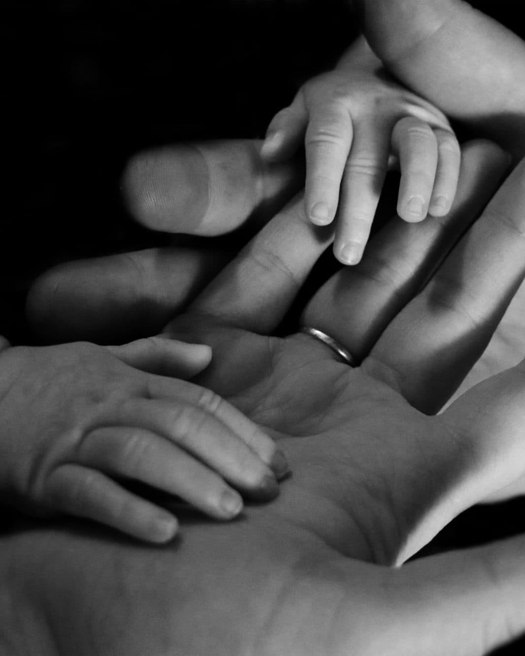 Una mano adulta abierta sostiene la mano de un niño pequeño y la de un bebé, con una alianza de boda visible, en blanco y negro sobre fondo oscuro