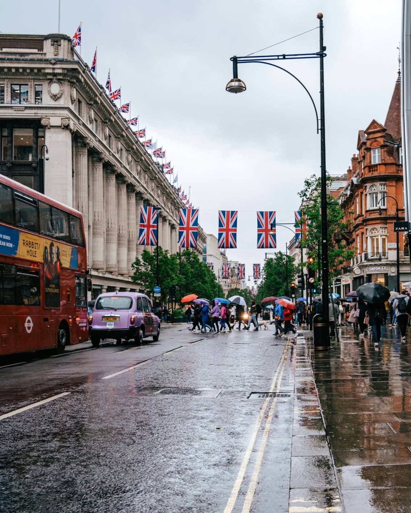 Una calle de Londres bajo la lluvia con un autobús rojo de dos pisos, un taxi, peatones cruzando con paraguas y banderas del Reino Unido tendidas sobre la calzada