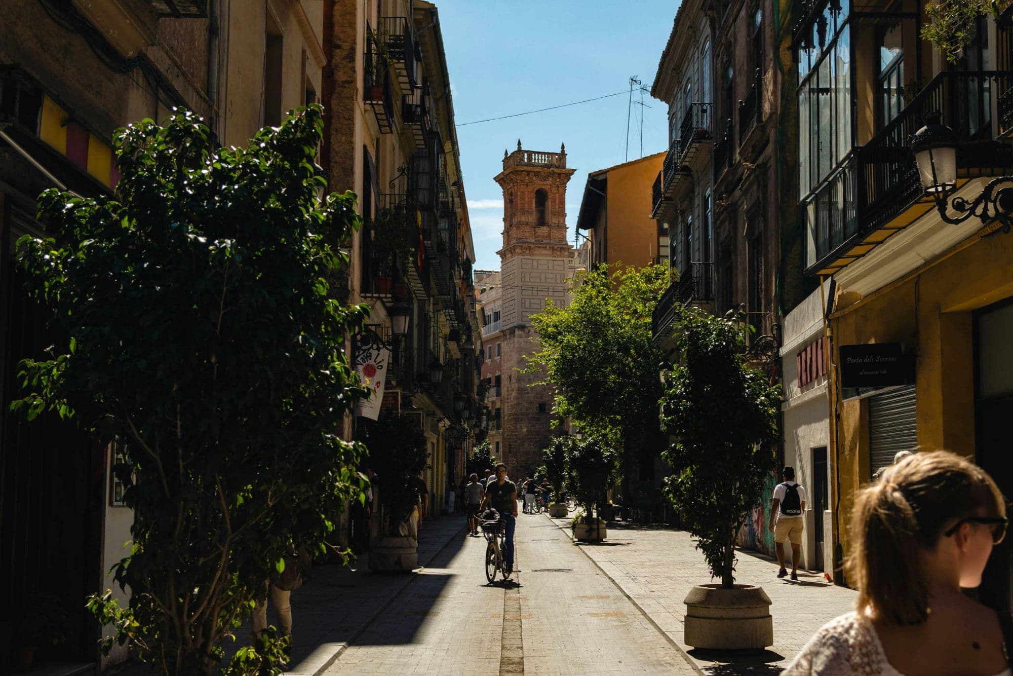 Una calle peatonal del centro histórico de Valencia en perspectiva central, con naranjos en macetas, un ciclista y peatones, y el remate de una torre barroca al fondo