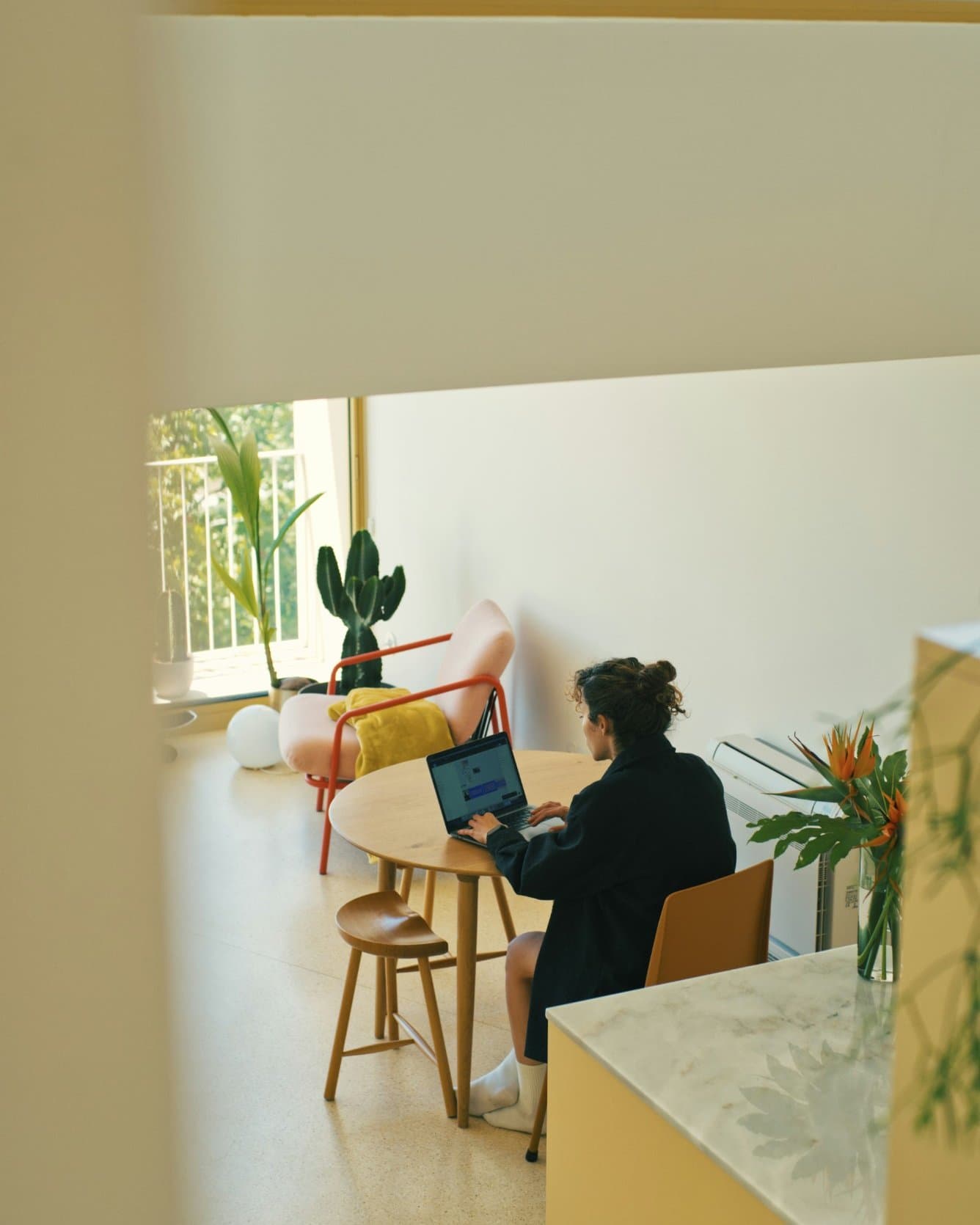 Una profesional trabaja con un portátil en una mesa redonda dentro de un interior luminoso, vista a través de un hueco arquitectónico con plantas y mobiliario contemporáneo