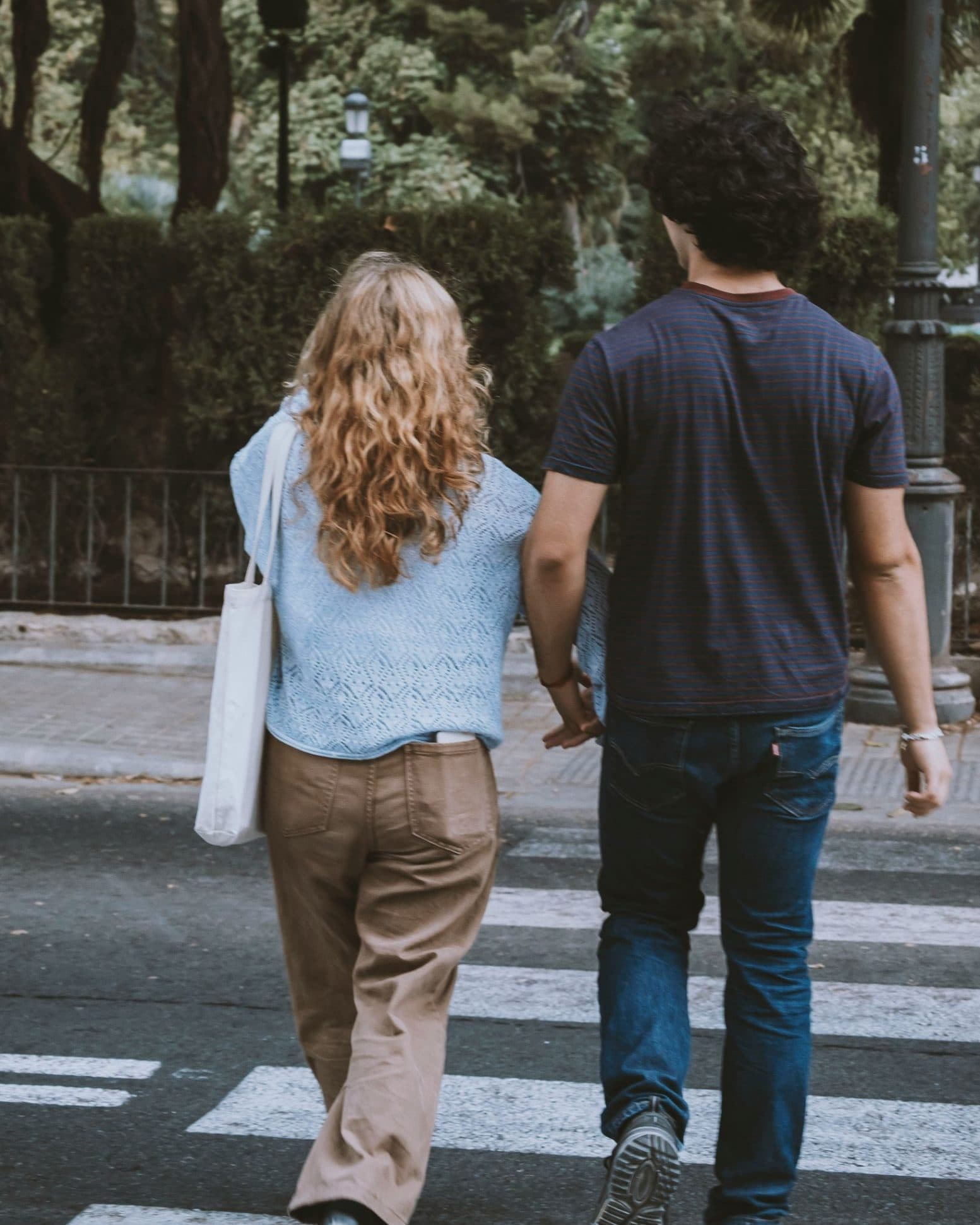 Una pareja camina de espaldas, cogida de la mano, cruzando un paso de peatones entre árboles y setos de una calle urbana